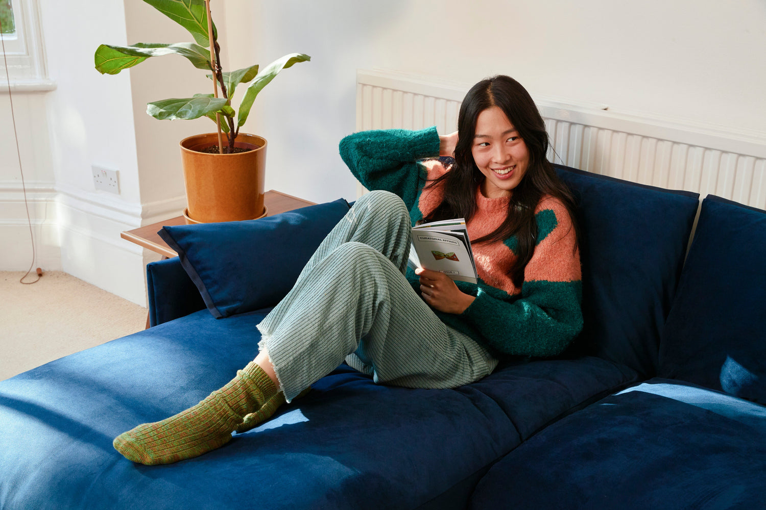 Woman relaxing on a deep blue velvet sofa in a bright living room with soft spring sunlight, reading a book beside indoor plants and warm wood accents.